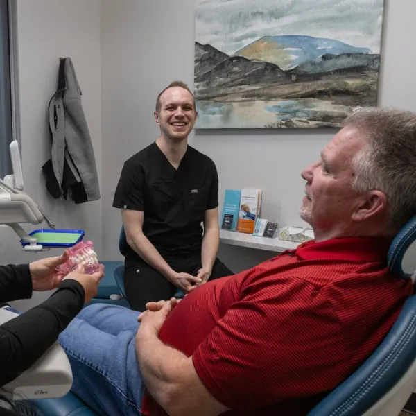 Dentist in black scrubs smiling while consulting an older male patient in a dental chair with dental molds nearby