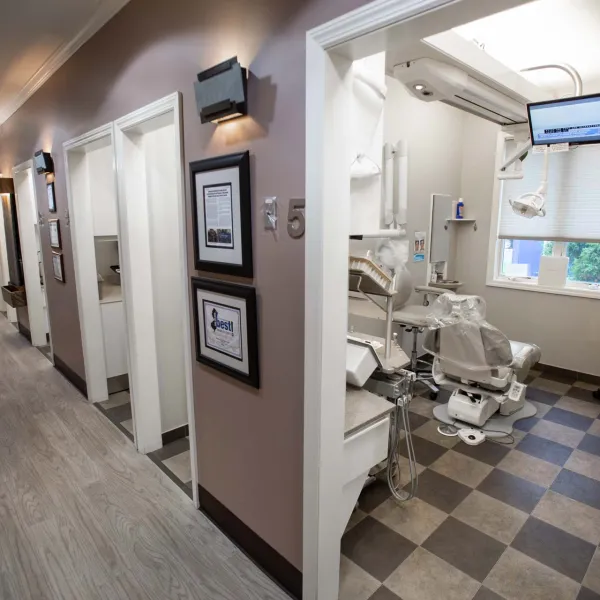 Bright dental clinic hallway with open patient rooms and modern dental equipment in one room visible on checkered floor.