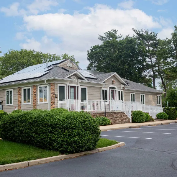 Single-story house with solar panels on the roof surrounded by greenery and an empty parking lot under a blue sky.