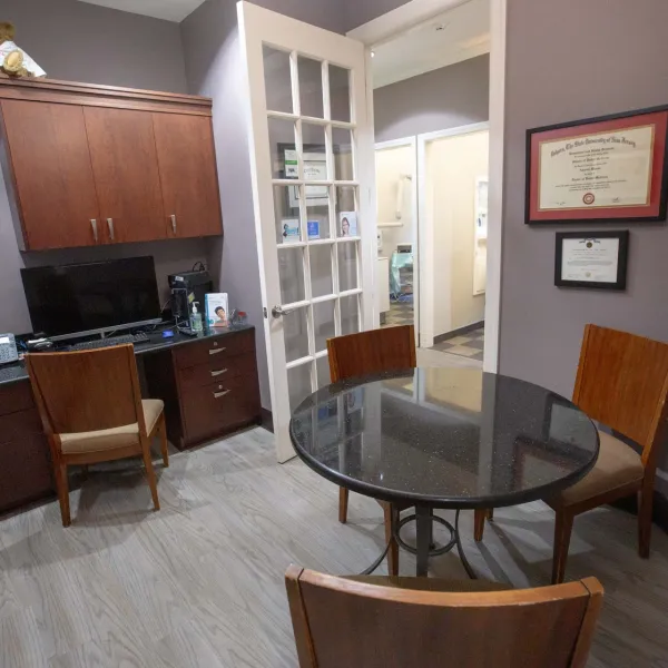 Small professional office with round table, three chairs, computer desk, certificates on gray walls, and wood cabinetry.