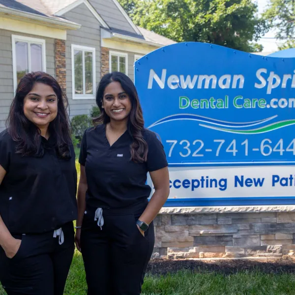Two smiling women in black scrubs stand outside Newman Springs Dental Care with a sign offering new patients.