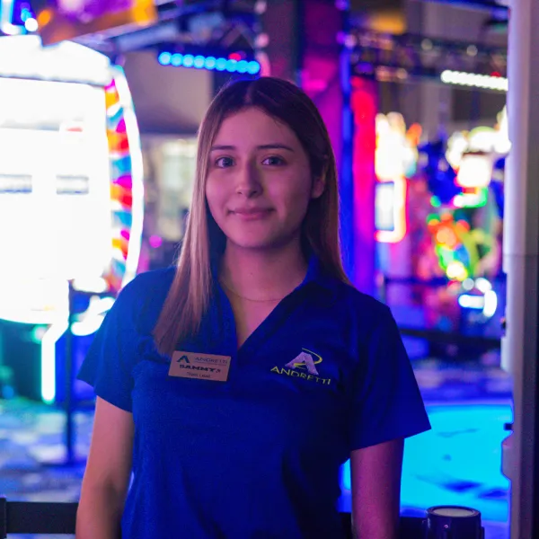 Smiling female staff member in blue Andretti uniform stands in colorful arcade with vibrant lights