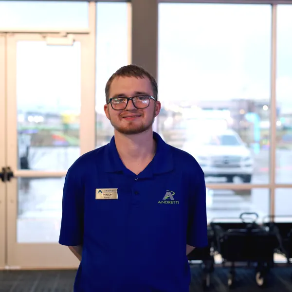 A young man in a blue polo shirt stands in a bright lobby area with large windows behind him.