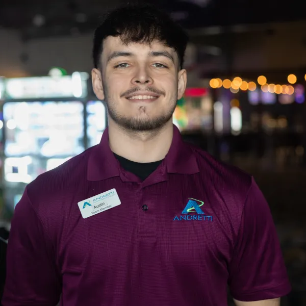 Smiling male staff member in an Andretti polo shirt, with arcade games blurred in the background.