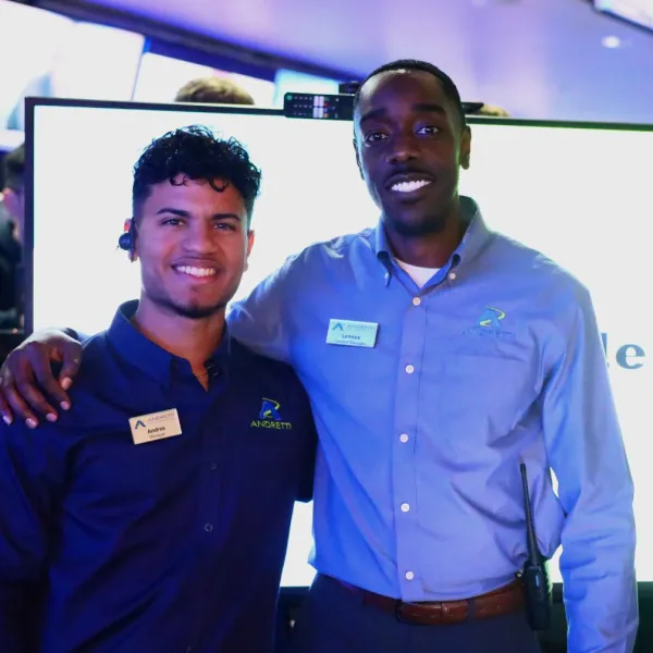 Two smiling male employees wearing name tags and company shirts pose in front of a digital display in a modern office.