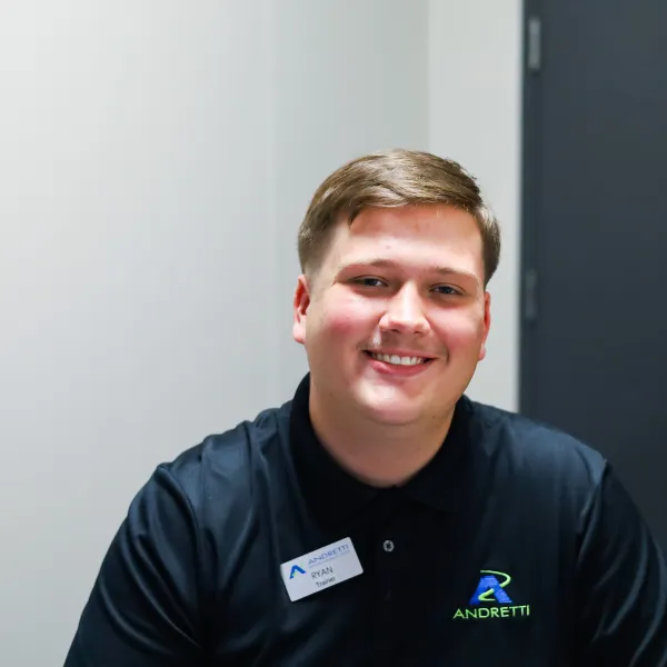 Smiling young man in a black polo shirt with Andretti logo, seated at a desk in a modern office.