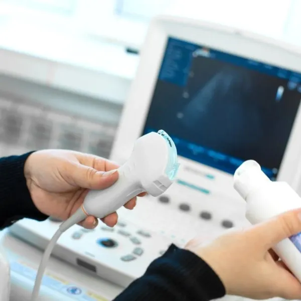Hands holding ultrasound probe and gel bottle in front of ultrasound machine monitor in medical setting.