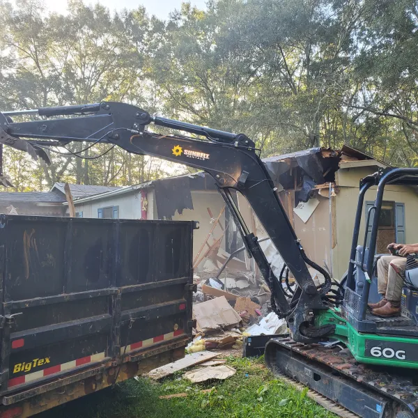 Man operating 60G excavator demolishing a house and loading debris into a BigTex dumpster outdoors