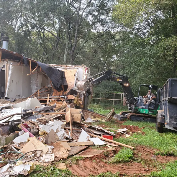 Excavator demolishing a wooden house surrounded by debris and trees in a green yard.
