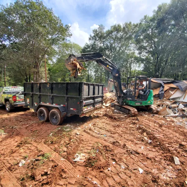 Mini excavator loading debris into a large trailer beside a pickup truck on a dirt lot during demolition work.