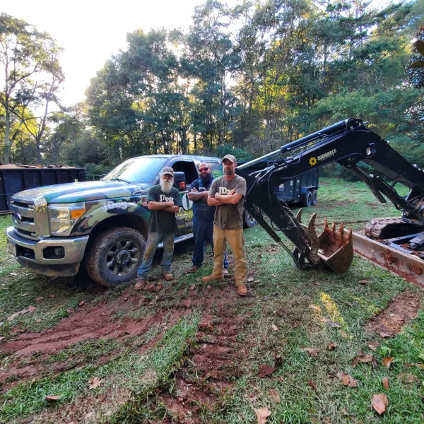 Three men standing in front of a camo truck and excavator in a wooded area with green grass and dirt tracks.