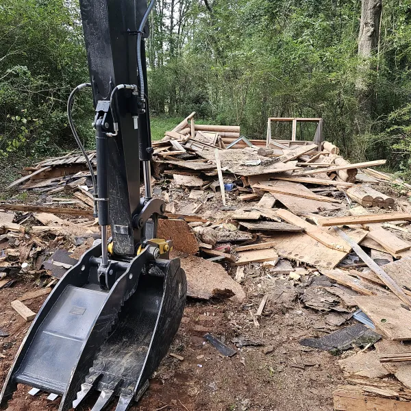 Excavator bucket in front of a large pile of wooden debris and construction waste in a forested area.