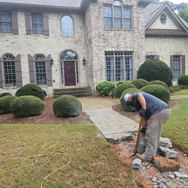 Worker breaking concrete sidewalk outside a large brick house with landscaping and shrubs.