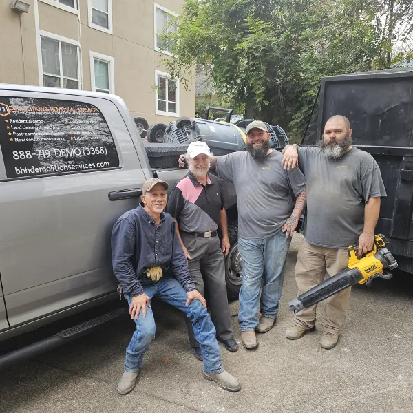 Four demolition workers stand and sit by a truck and dumpster, posing with tools and smiling outside a building.
