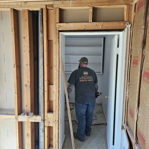 Man in construction shirt working inside partially demolished interior with exposed wooden framing and insulation.