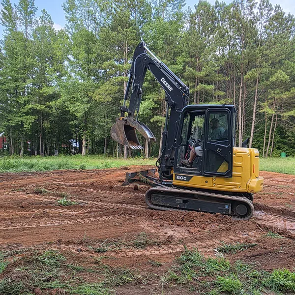 Yellow and black John Deere excavator digging on red dirt clearing surrounded by green trees under blue sky.