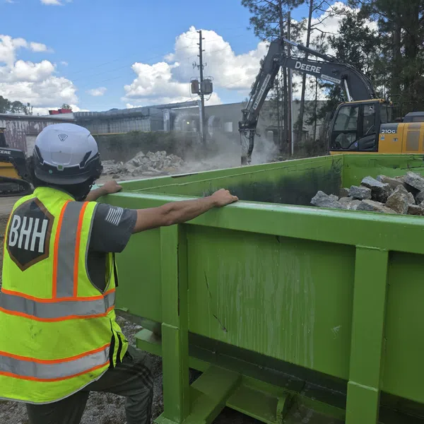 Construction worker in safety gear overseeing debris being loaded into a green dumpster with heavy machinery in background