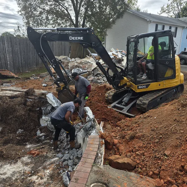 Construction workers operate a John Deere excavator breaking up concrete near a residential house.