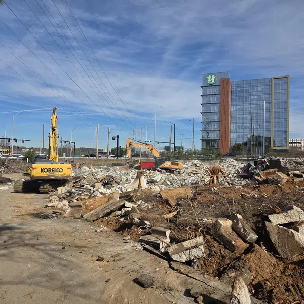 Construction site with excavators clearing rubble and debris near modern glass office buildings under a blue sky.