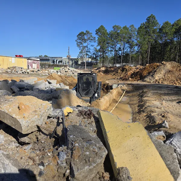 Construction site with excavator digging soil trench amid broken concrete slabs and dirt piles under clear blue sky.