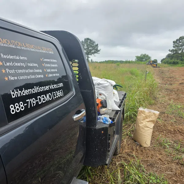 Black demolition service truck parked by a partly cleared grassy field with a driver in the cab on a cloudy day.