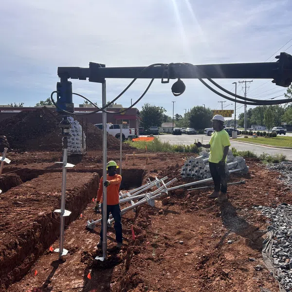 Construction workers operating machinery to install metal poles at a dirt site near a road on a sunny day.