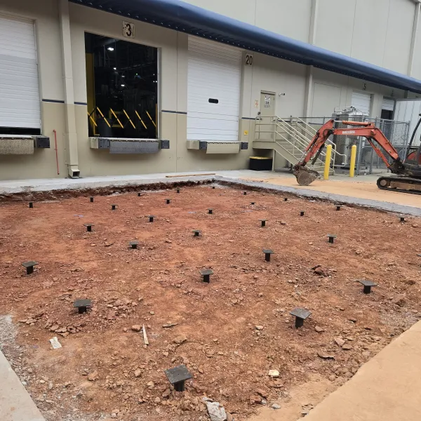 Construction site with helical support in front of warehouse loading docks and excavator.