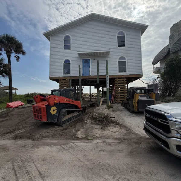 Two-story house elevated on stilts with construction equipment and a pickup truck nearby under a cloudy sky.