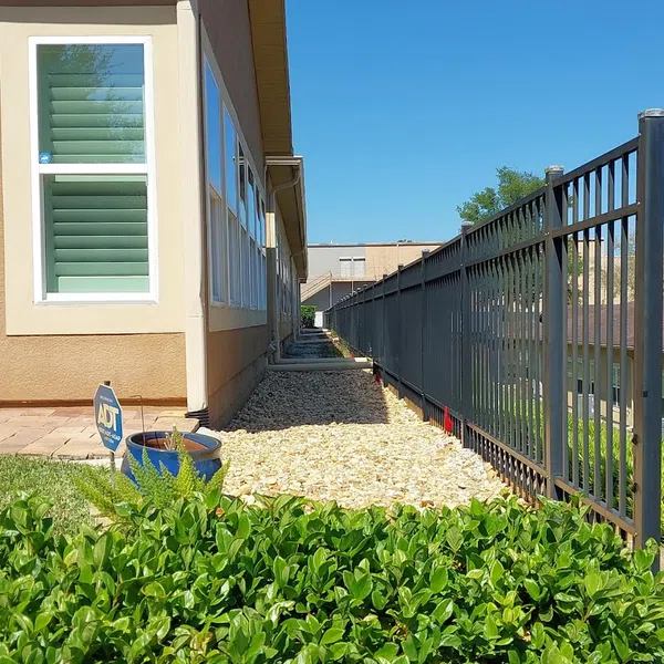 Side view of a beige house with white windows, black metal fence, gravel path, and green shrubbery under a clear blue sky