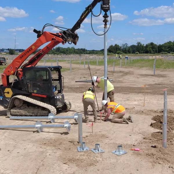 Construction workers install metal posts using an excavator in a large open field under a blue sky.