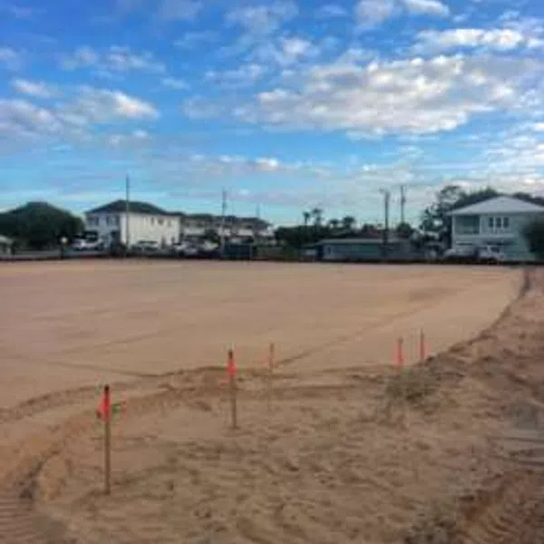 Construction site with leveled dirt ground marked by stakes and houses in the background under a blue sky.