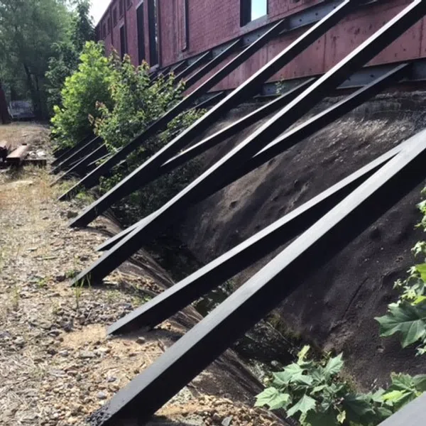 Steel braces supporting an old brick building with overgrown vegetation along the ground and wall.