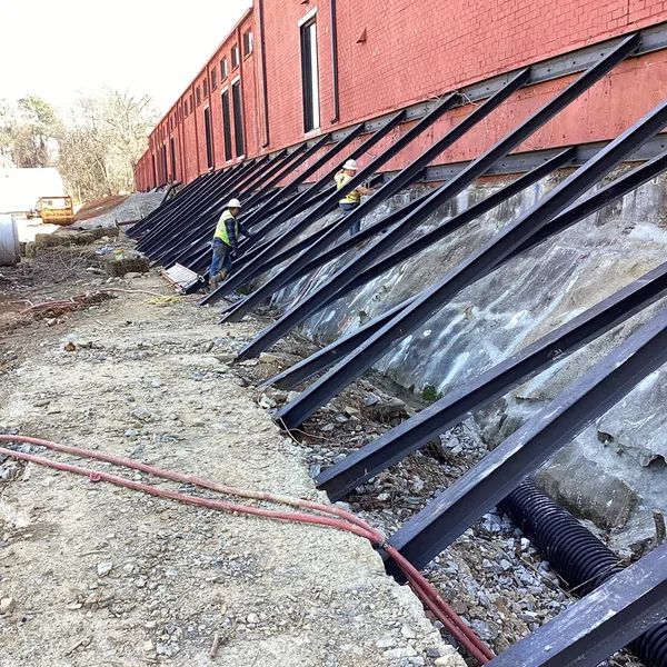 Construction workers install steel braces supporting a red brick building's foundation on a construction site.