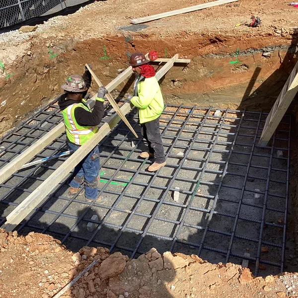 Construction workers installing wooden planks over reinforced steel grid in a deep foundation trench at a building site.