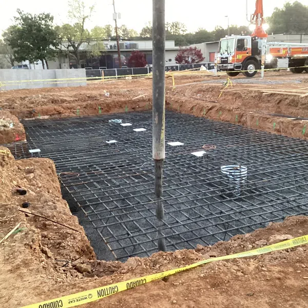Concrete pouring with rebar foundation at a construction site surrounded by caution tape and construction vehicles.