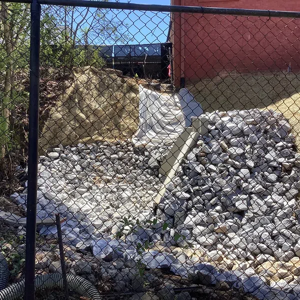 Rock-lined drainage channel with white fabric on a slope, fenced off near a brick building under blue sky.