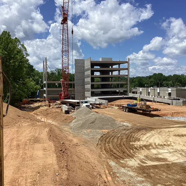 Construction site with heavy machinery, crane, dirt piles, and a multi-story concrete building structure under a blue sky.