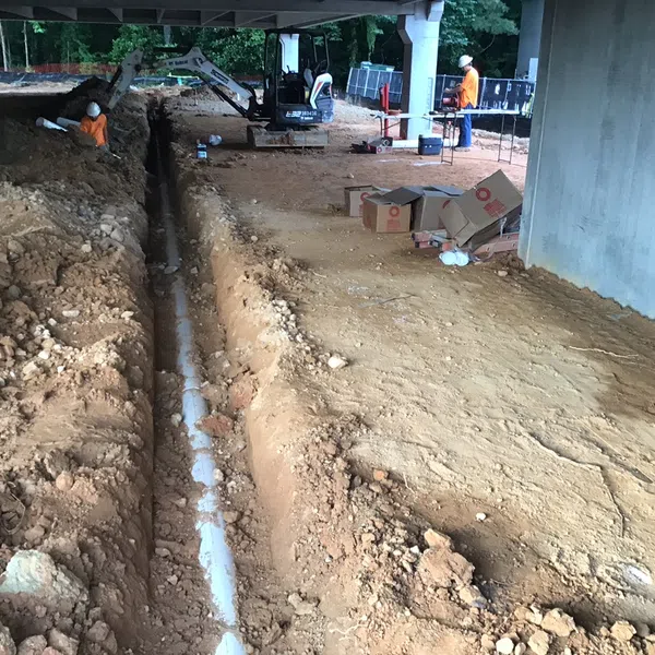 Construction site trench with workers installing underground white PVC piping below a bridge structure.