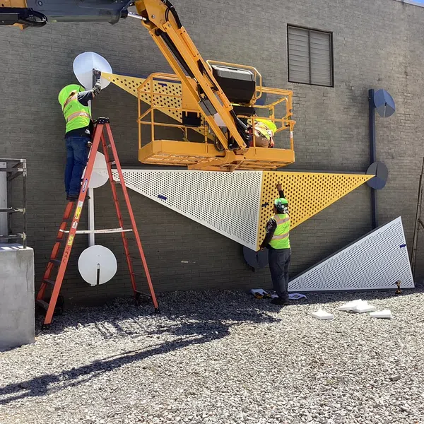 Workers installing yellow and white perforated metal panels on a building exterior using a lift and ladder