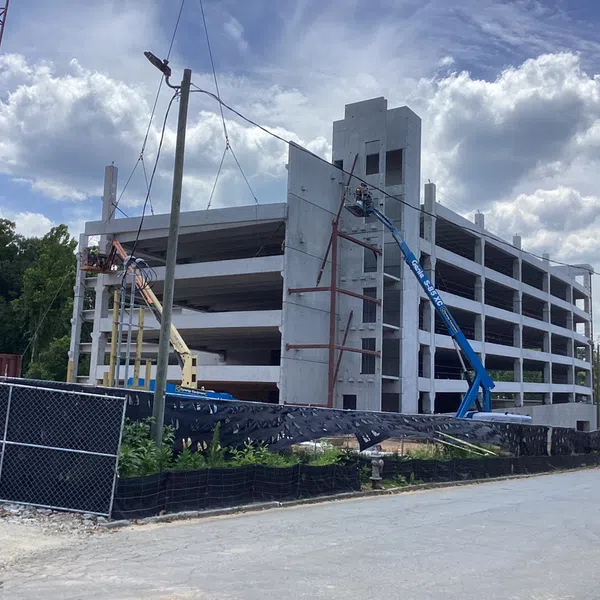 Multi-story concrete parking garage under construction with cranes, cherry pickers, and construction fencing outside.