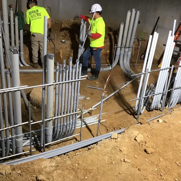 Construction workers installing electrical conduit pipes underground at a building site with safety gear.