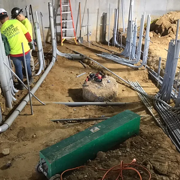 Two electricians working on underground electrical conduit installation in a construction site basement with tools and cables.