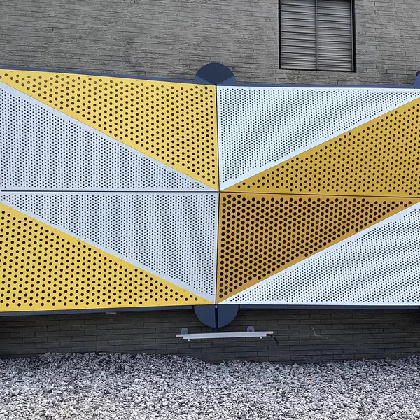 Yellow and white perforated metal panels arranged in geometric pattern mounted on a brick wall above gravel.