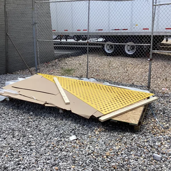 Piles of construction materials including yellow perforated board and wooden planks on gravel near chain-link fence.