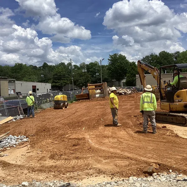 Construction workers in safety vests operating heavy machinery on a cleared dirt site under a blue sky with clouds.
