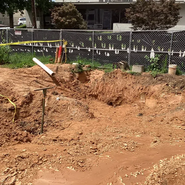 Excavated construction site with exposed dirt, a white pipe, and a chain-link fence surrounding the area on a sunny day