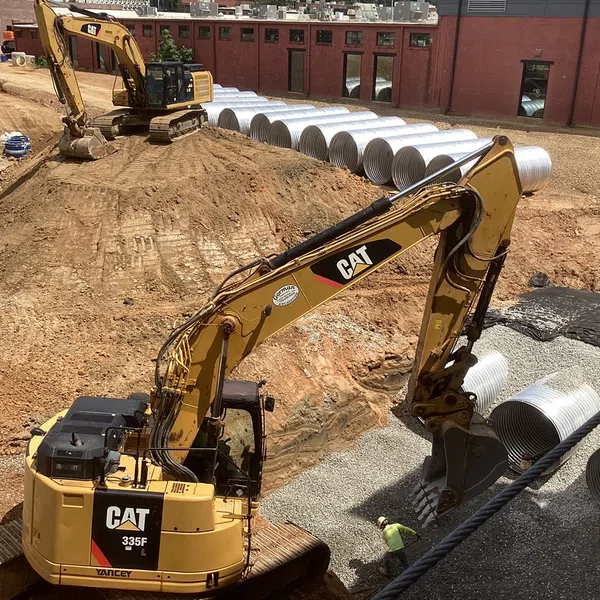 Two CAT excavators working on construction site with large metal drainage pipes and gravel base.