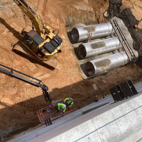 Construction site with excavator, workers on lift, and large metal drainage pipes on dirt ground