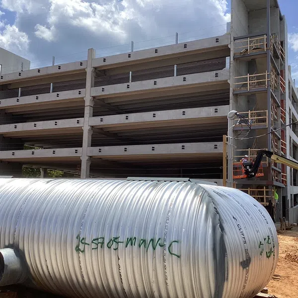 Multi-level concrete parking garage under construction with large cylindrical metal drainage pipe in foreground under blue sky.