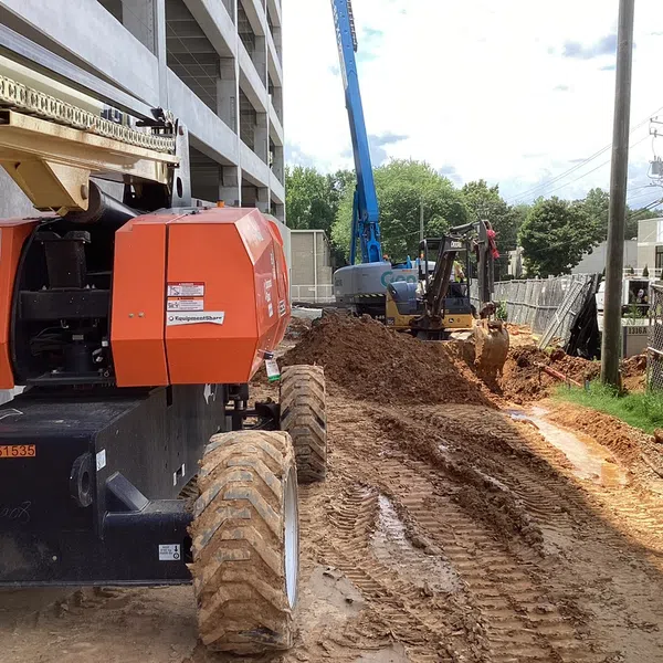Construction site with mud, heavy equipment including an orange boom lift and excavator near a building and fence.